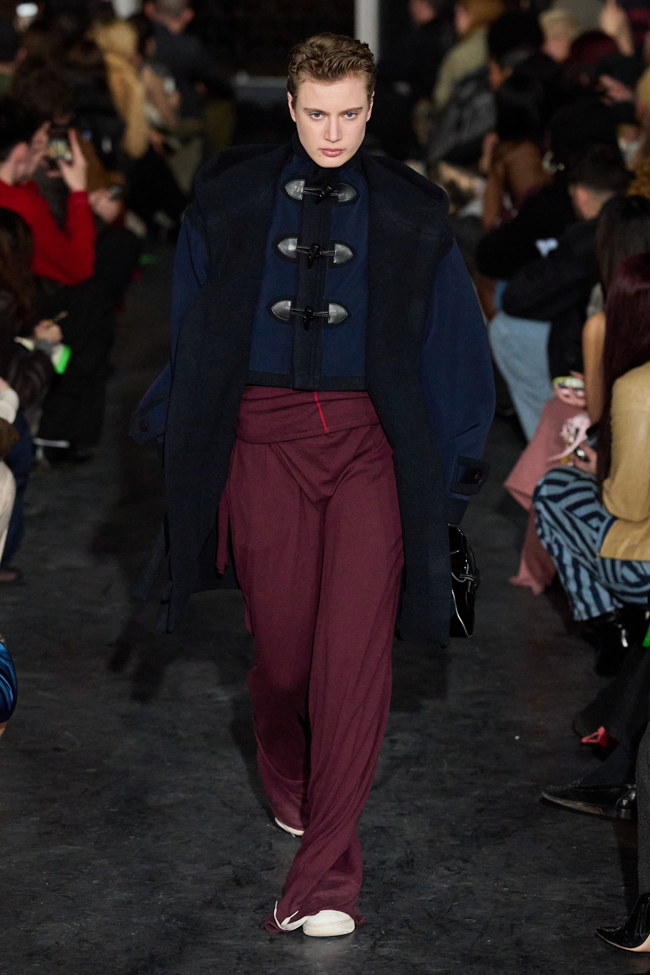 Модель в тёмном пальто и бордовых брюках, создающих эффектный контраст с верхом, уверенно шагает по подиуму