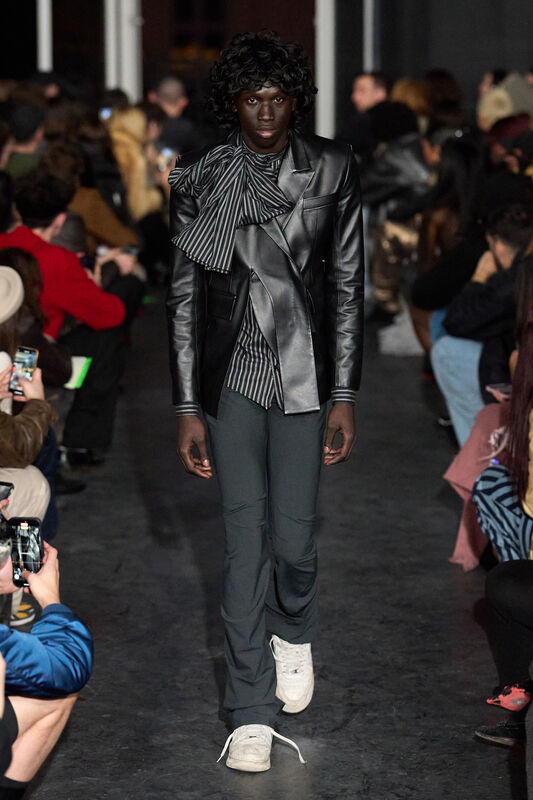 Модель в черном кожаном пиджаке с полосатым элементом, сочетает строгий стиль с игривыми деталями, уверенно шагая по подиуму