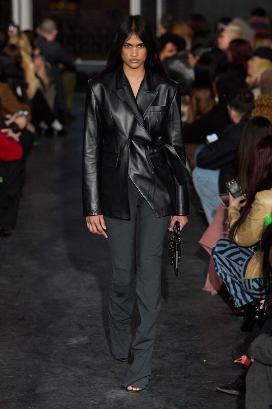Кожаный пиджак с поясом и брюки в тон, создают стильный образ, подчеркивающий силуэт модели на фоне зрителей