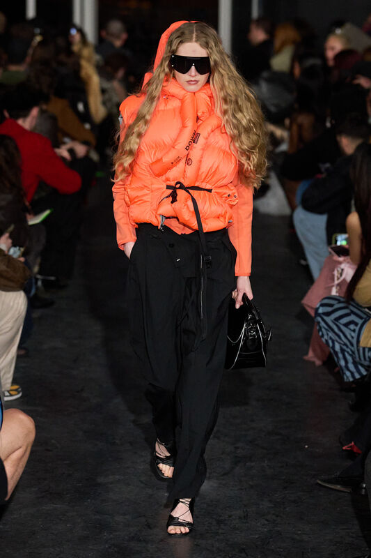 Ярко-оранжевый пуховик с капюшоном и черными широкими брюками, стильный аксессуар — черная сумка