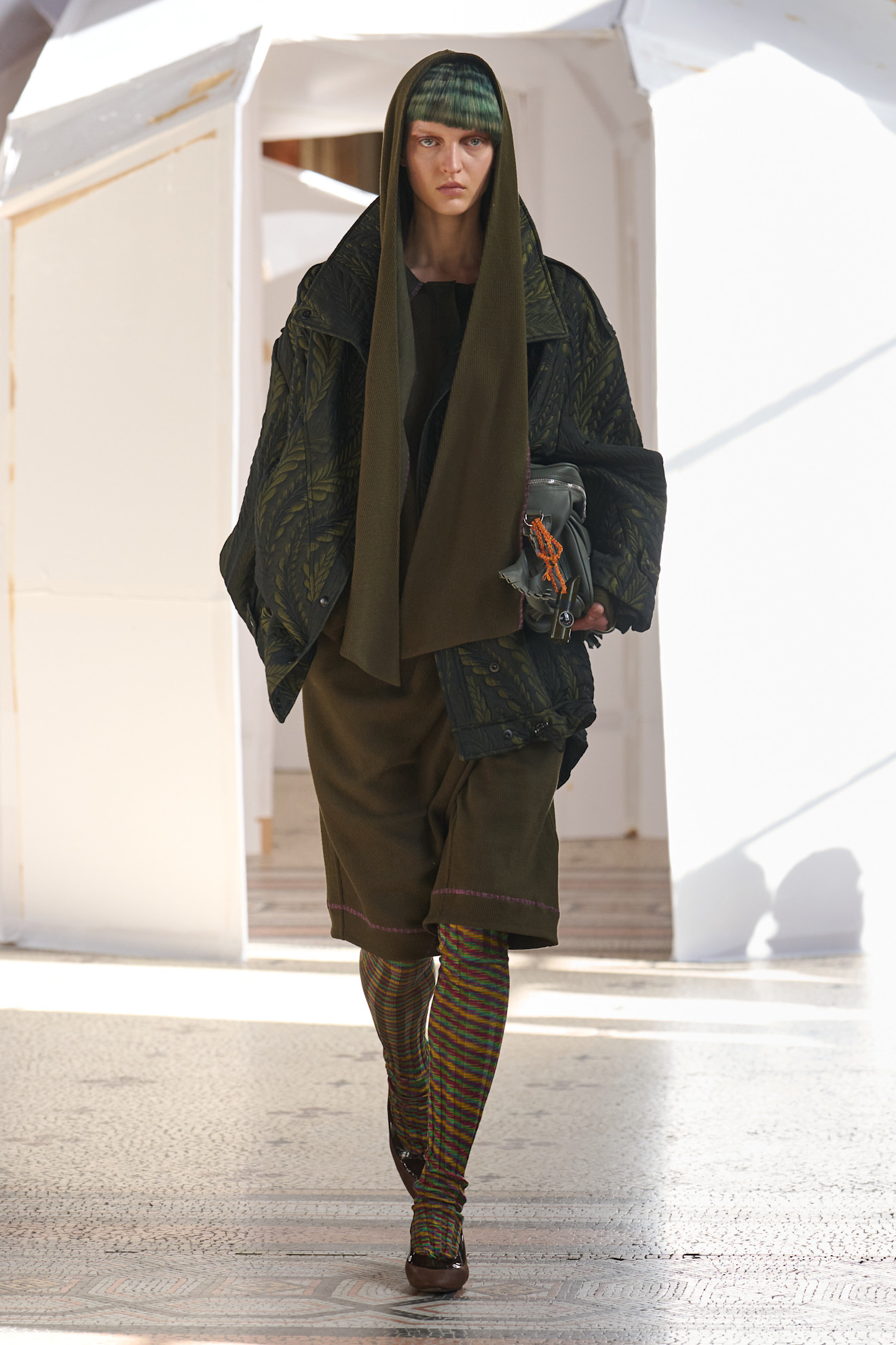 Модель в многослойном образе с капюшоном и яркими колготками, сочетает уют и современный стиль с акцентом на детали