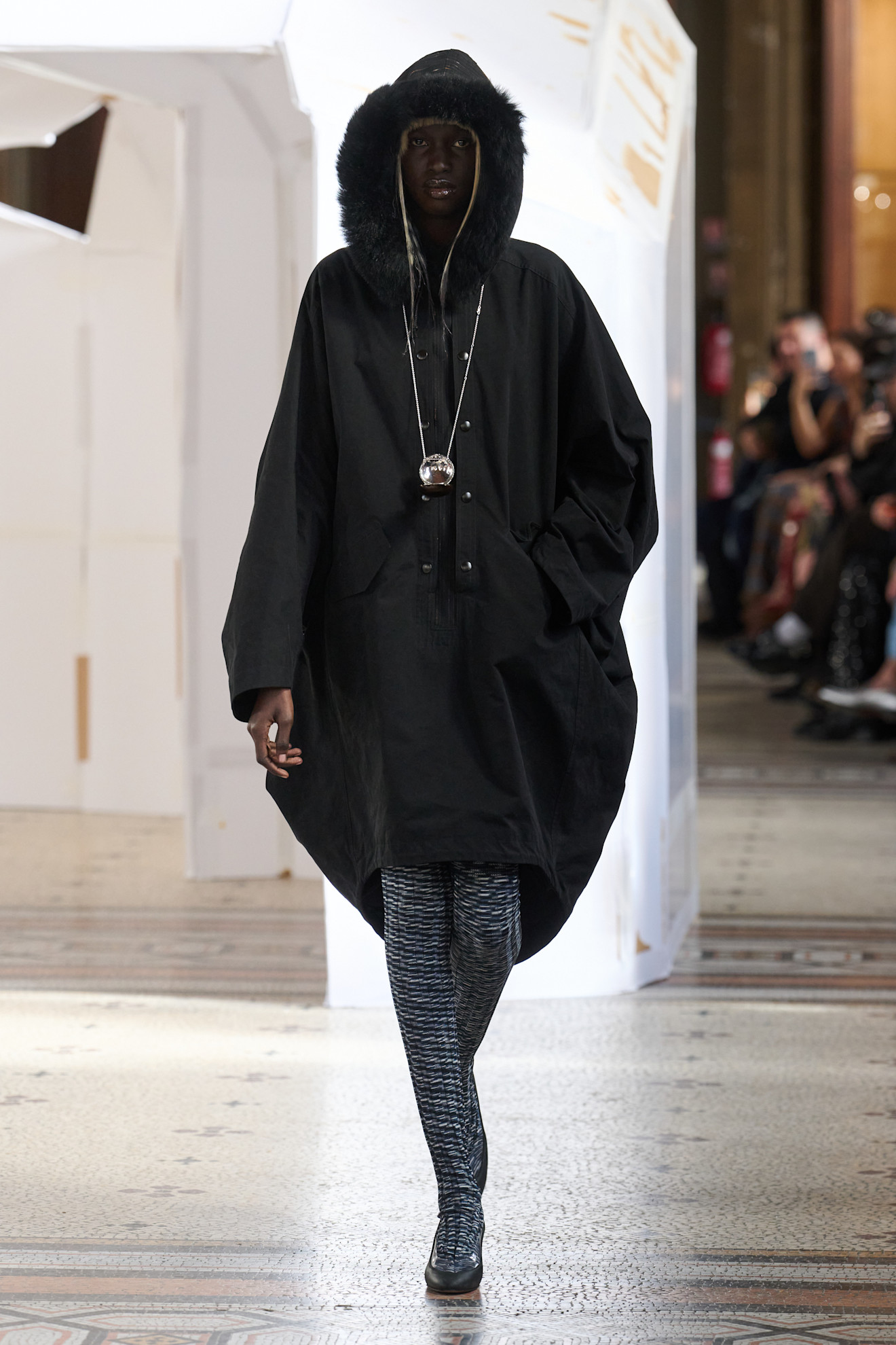 Объемная черная куртка с меховым капюшоном, дополненная стильной подвеской и узкими колготками с узором