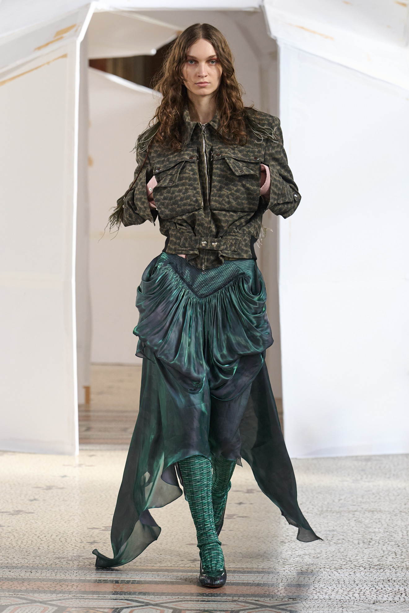 Модель в жакете с камуфляжным принтом и струящемся зелёном юбке, создающем эффект движения, на фоне минималистичного пространства