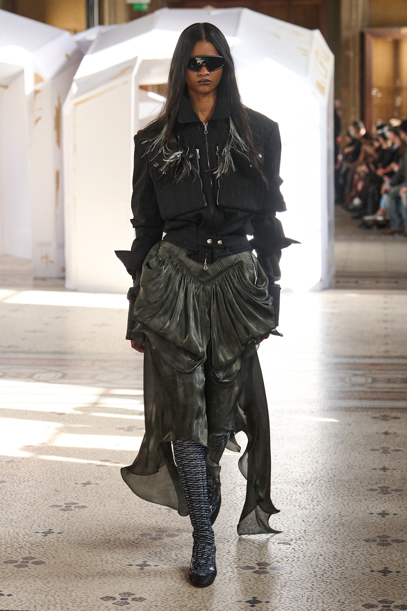 Модель в черном наряде с металлическими деталями и объемной юбкой, акцент на текстуре и контрасте с ярким светом