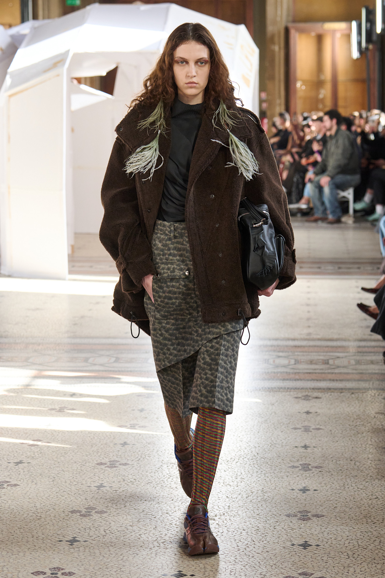 Объемный коричневый пиджак с зелеными акцентами и асимметричная юбка в клетку, создающие уютный, но современный образ