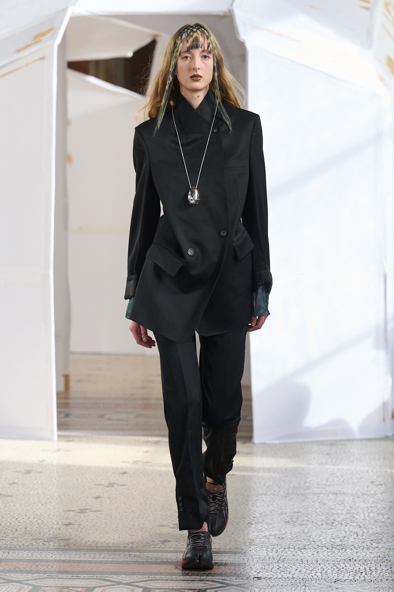 Строгий черный костюм с асимметричным жакетом и кроссовками, дополненный длинной цепочкой с кулоном