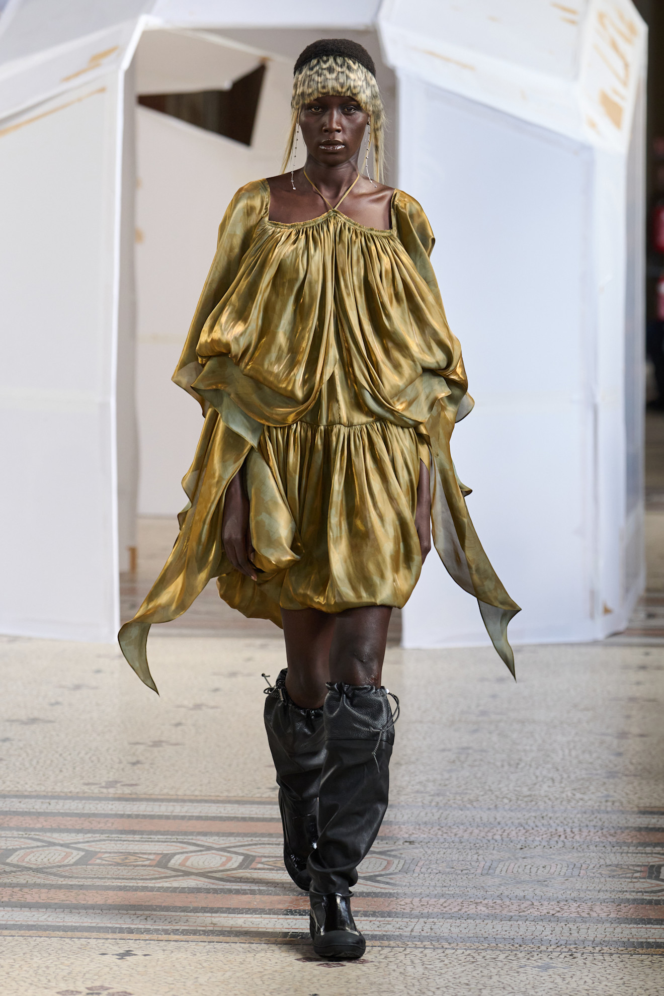 Объемное платье золотистого оттенка с драпировками, дополненное высокими черными сапогами, создает эффектный контраст.