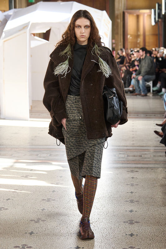 Объемный коричневый пиджак с зелеными акцентами и асимметричная юбка в клетку, создающие уютный, но современный образ
