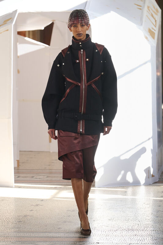 Модель в черной куртке с бордовыми вставками и асимметричной юбкой, создающей динамичный силуэт