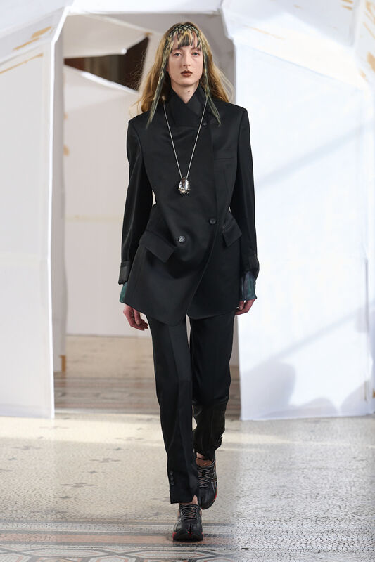 Строгий черный костюм с асимметричным жакетом и кроссовками, дополненный длинной цепочкой с кулоном