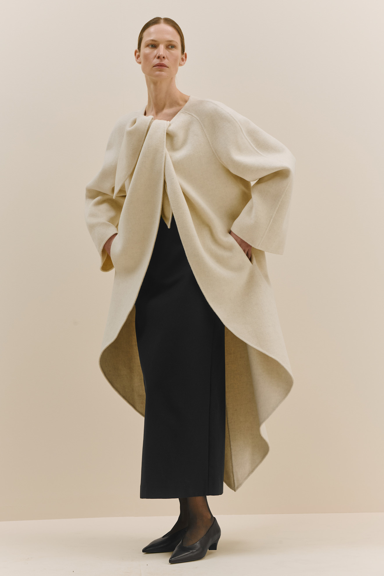 Стильный бежевый плащ с крупным бантом, дополненный черной юбкой, придаёт образу современный и утончённый вид.