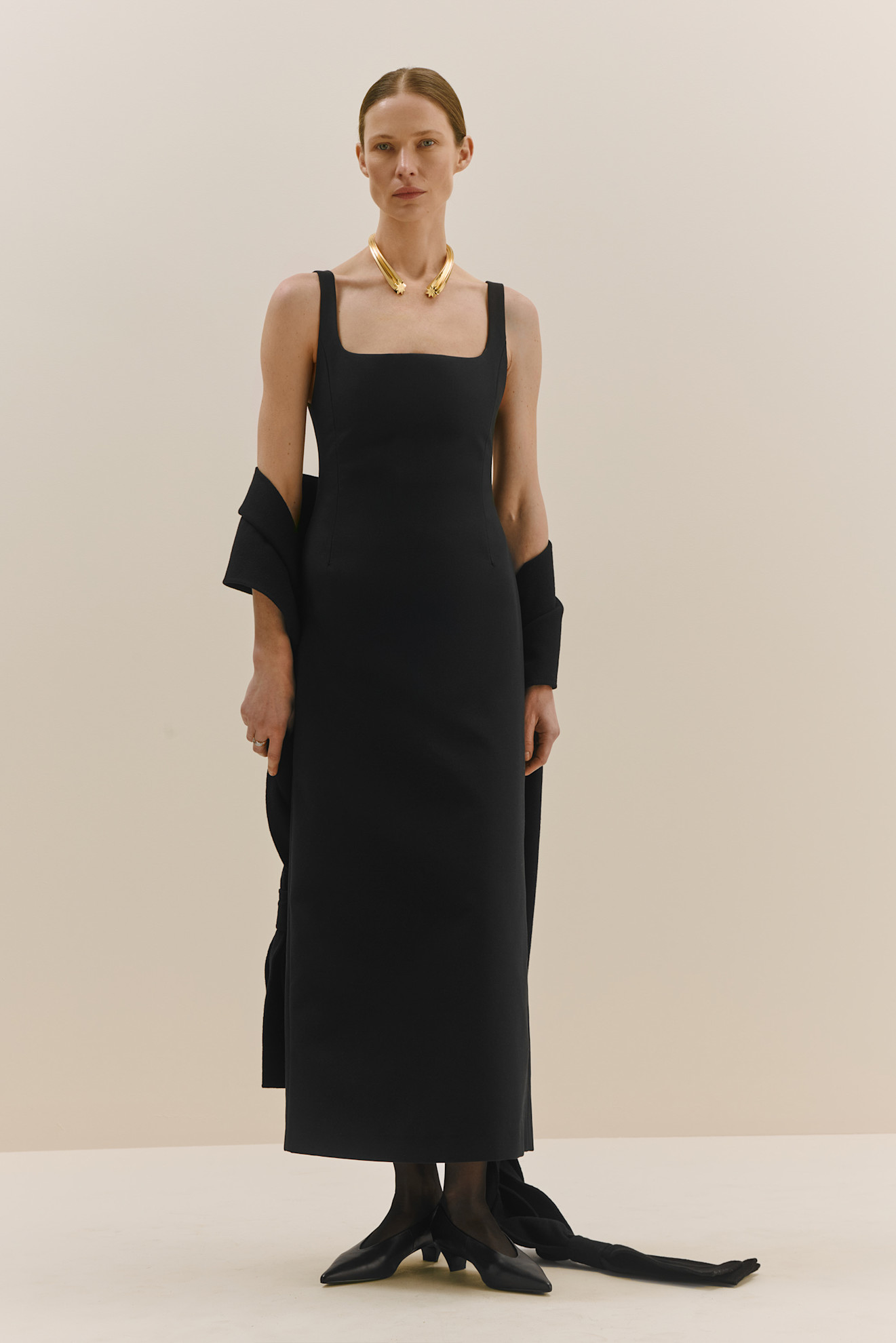 Минималистичное черное платье с квадратным вырезом, дополненное золотым ожерельем, излучает современный шик и уверенность