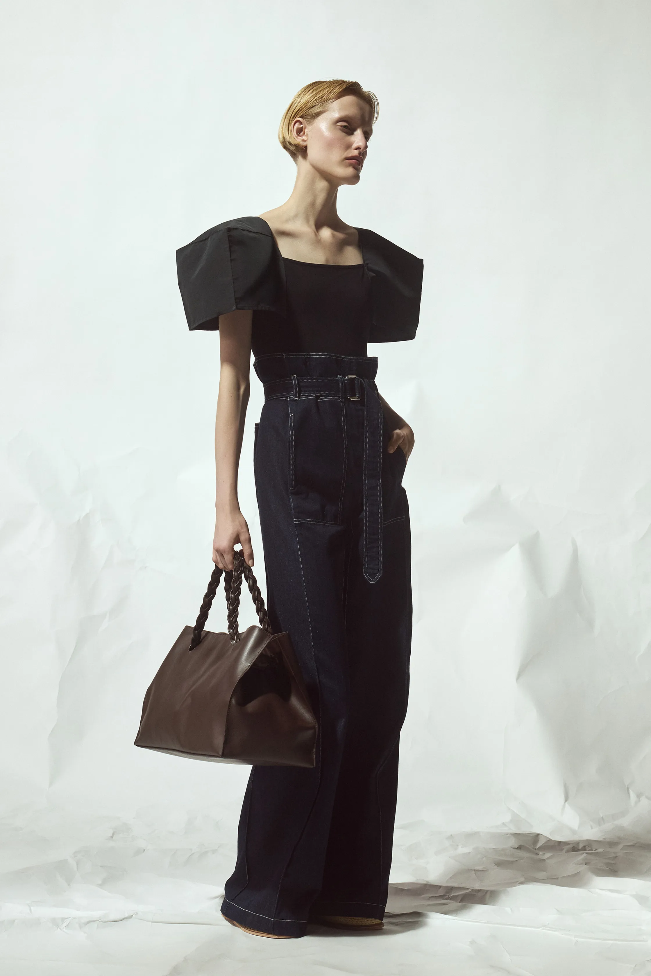 Черный топ с объемными рукавами и высокие джинсы с поясом, гармонично сочетающиеся с коричневой сумкой, создающей стильный акцент