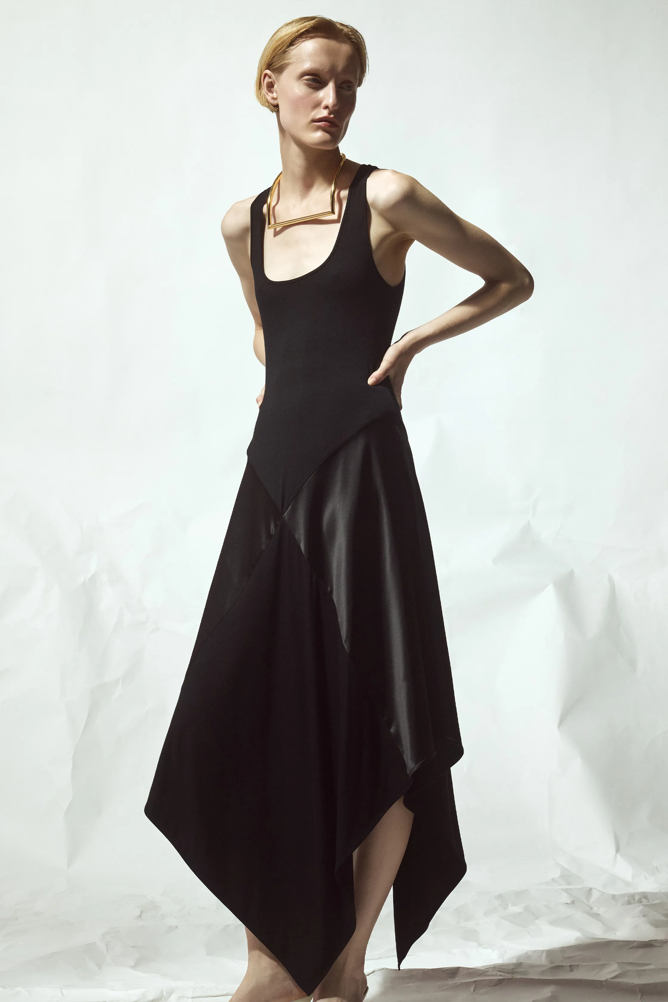 Минималистичное черное платье с асимметричным подолом и глубоким вырезом, дополненное золотым ожерельем