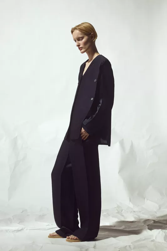 Объемный черный костюм с глубоким вырезом и широкими брюками, создающий эффектный контраст с минималистичным фоном