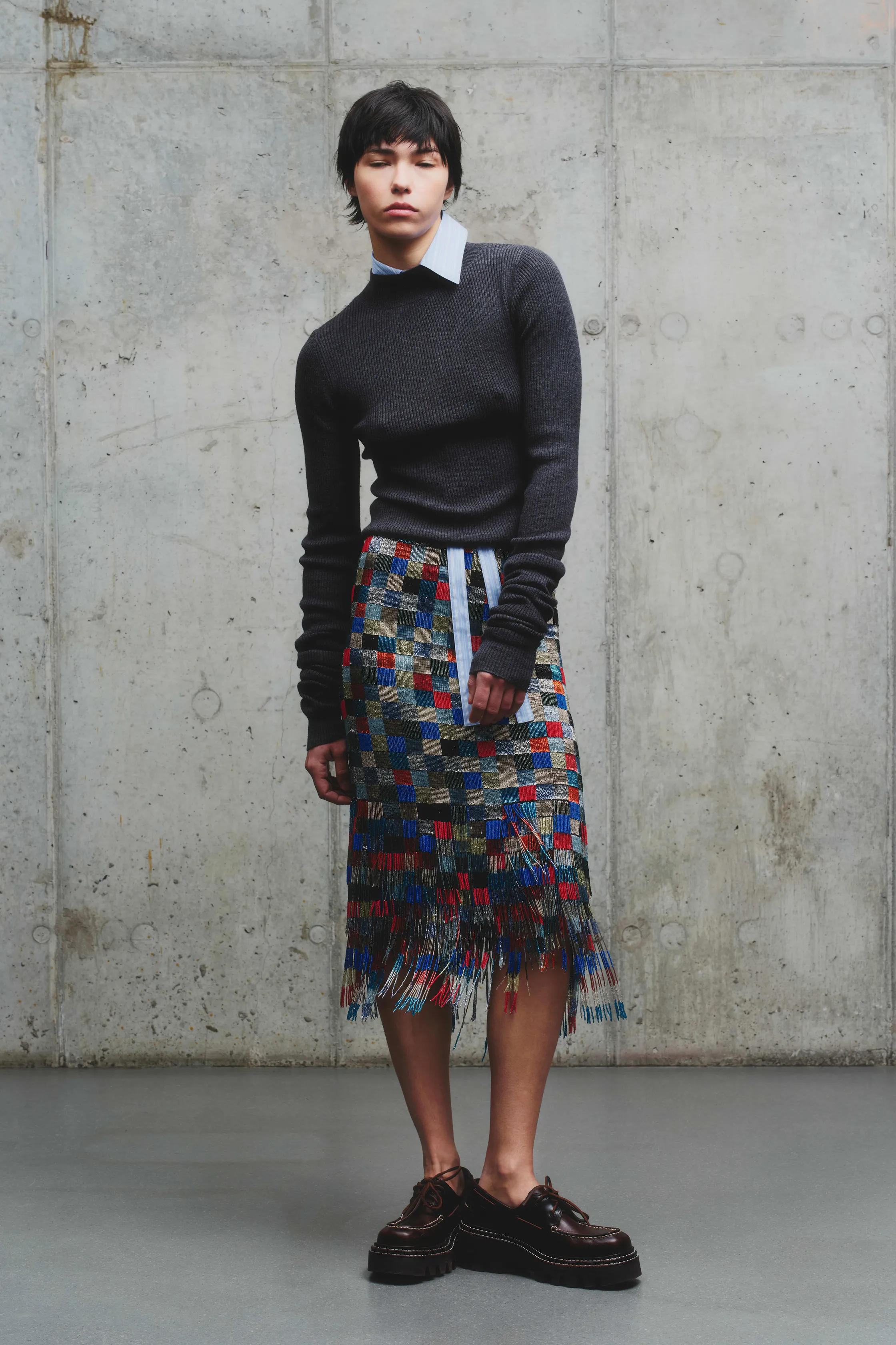 Теплый серый свитер в сочетании с яркой юбкой в клетку с бахромой, создающей динамичный образ на фоне бетонной стены