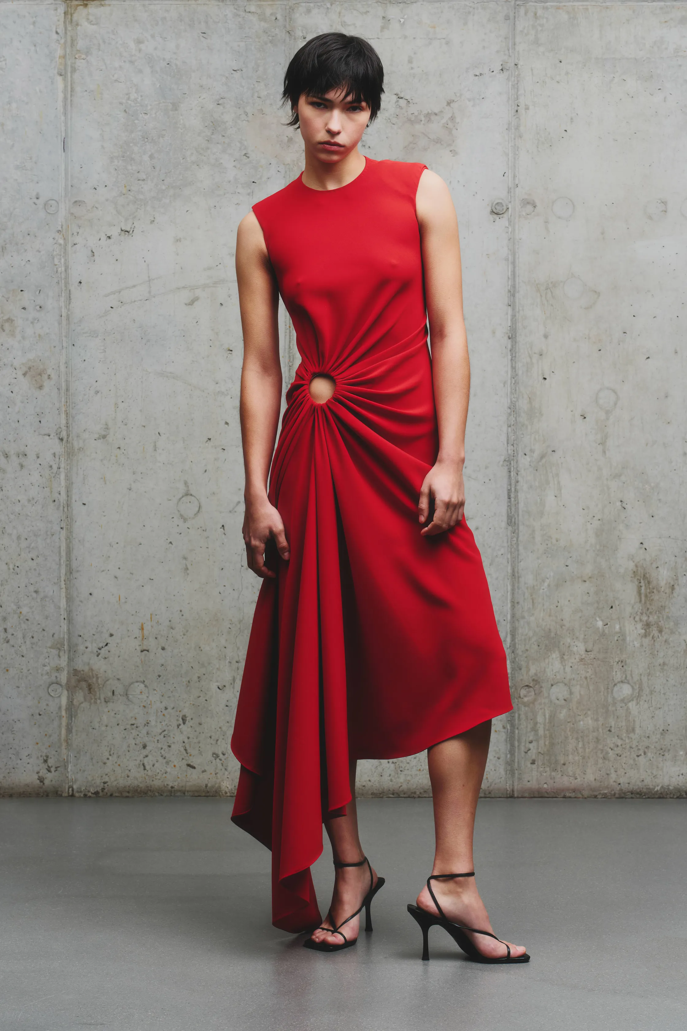 Элегантное красное платье с асимметричным кроем и круглым вырезом, подчеркивающее силуэт, на фоне холодного бетона