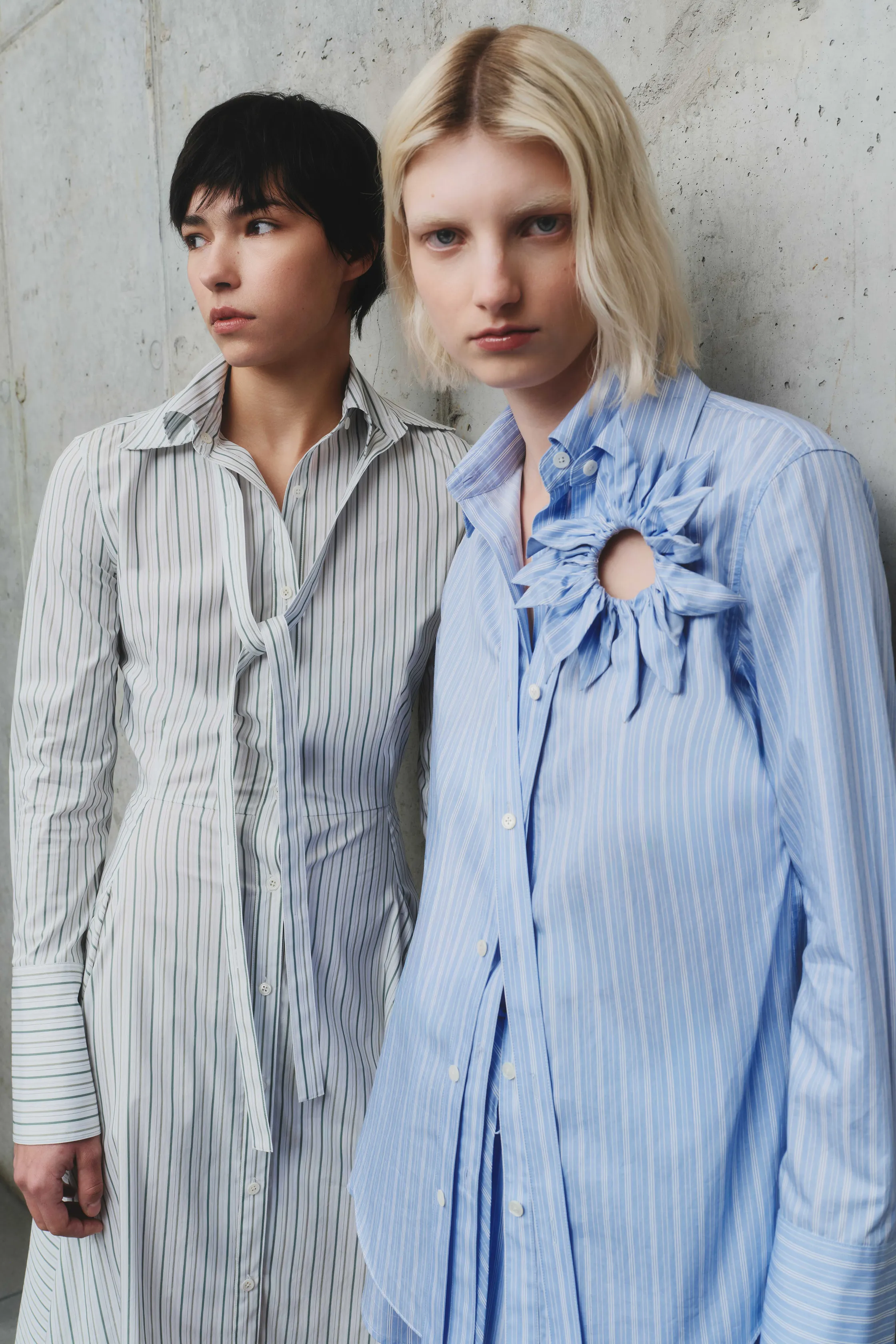 Две модели в полосатых рубашках, одна с рюшами, другая с завязкой, создают контрастные образы на фоне текстурной стены