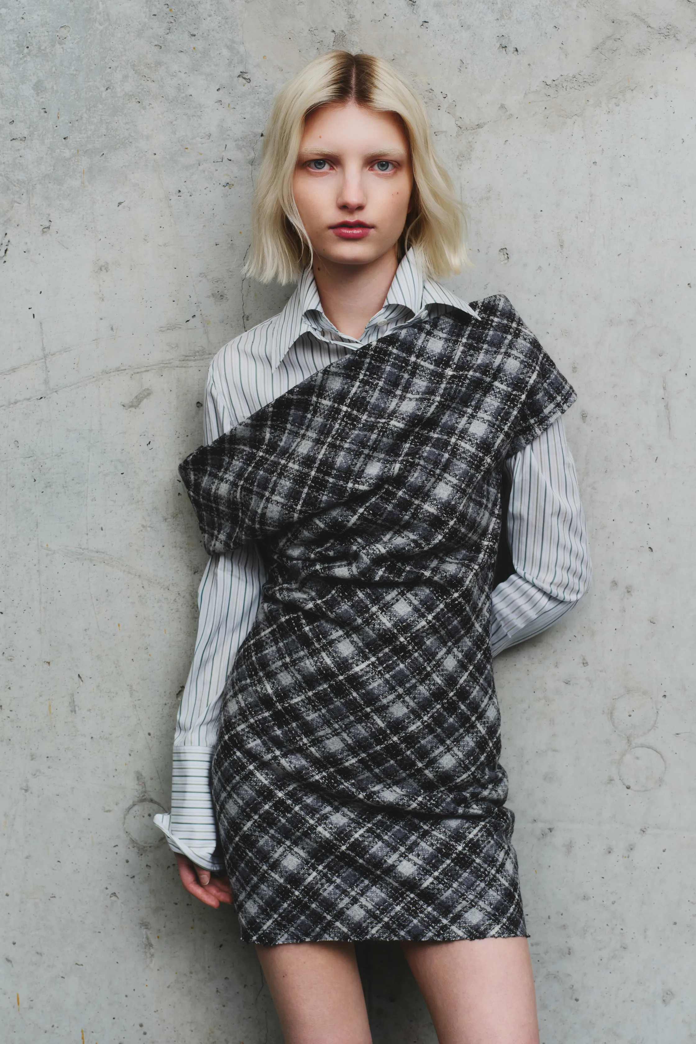 Модель в черно-серой клетчатой мини-юбке и полосатой рубашке, изящно обрамляющей фигуру, на фоне бетонной стены