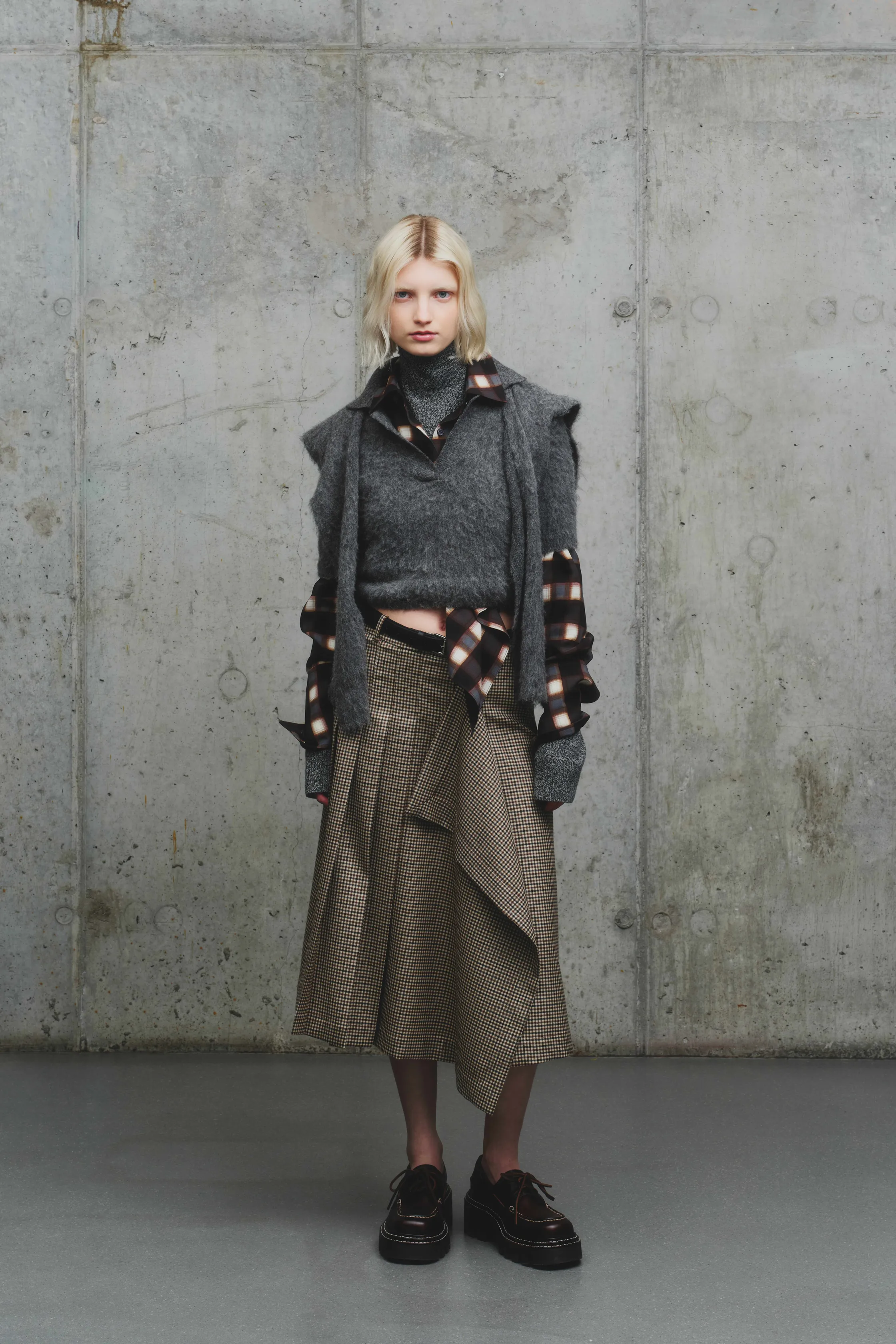 Стильный образ с серым свитером и клетчатой юбкой, дополненный многослойностью и текстурой, на фоне индустриального интерьера