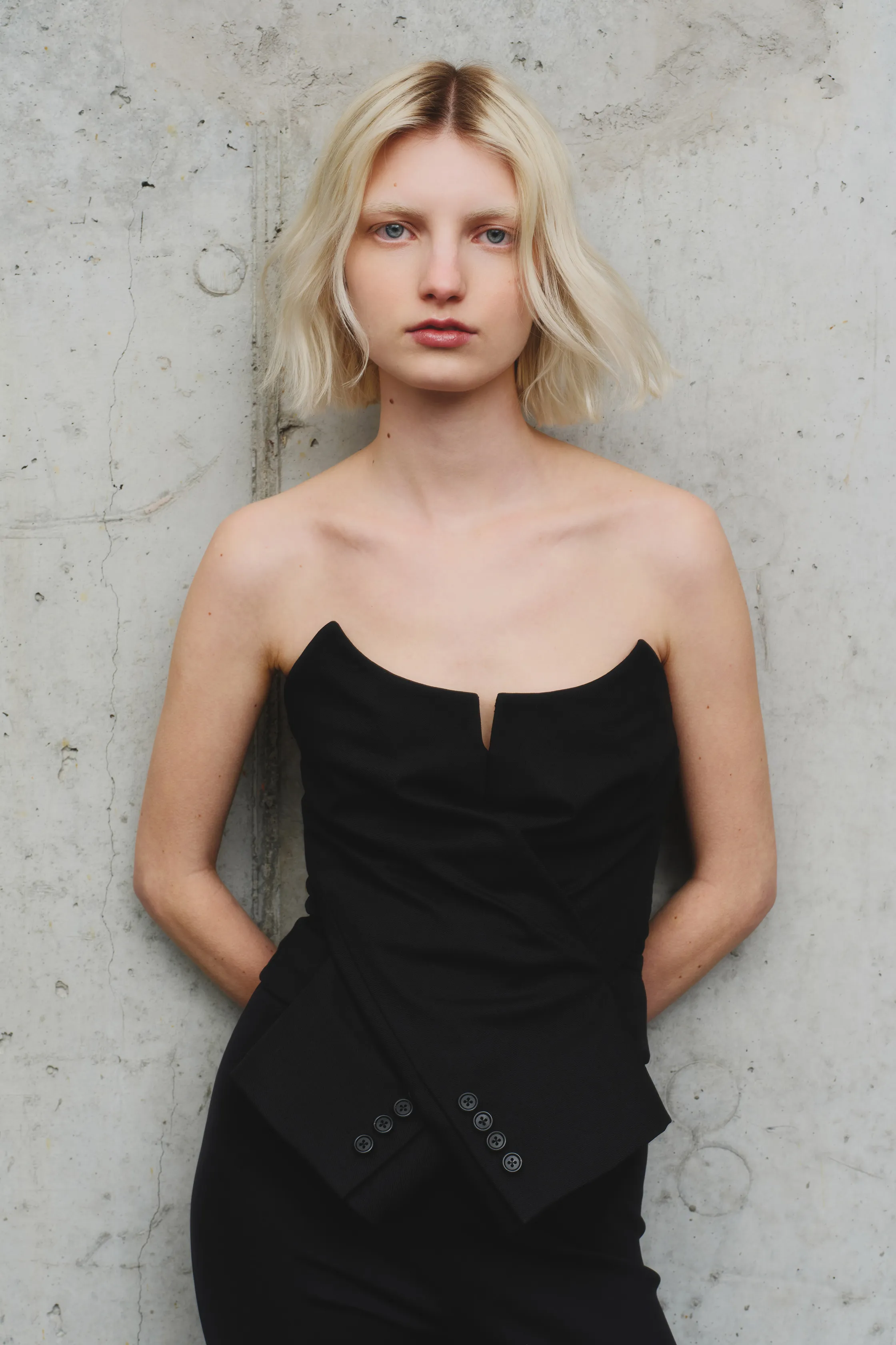 Черный топ с глубоким вырезом и асимметричным кроем, подчеркивающий стройную фигуру модели на фоне бетонной стены