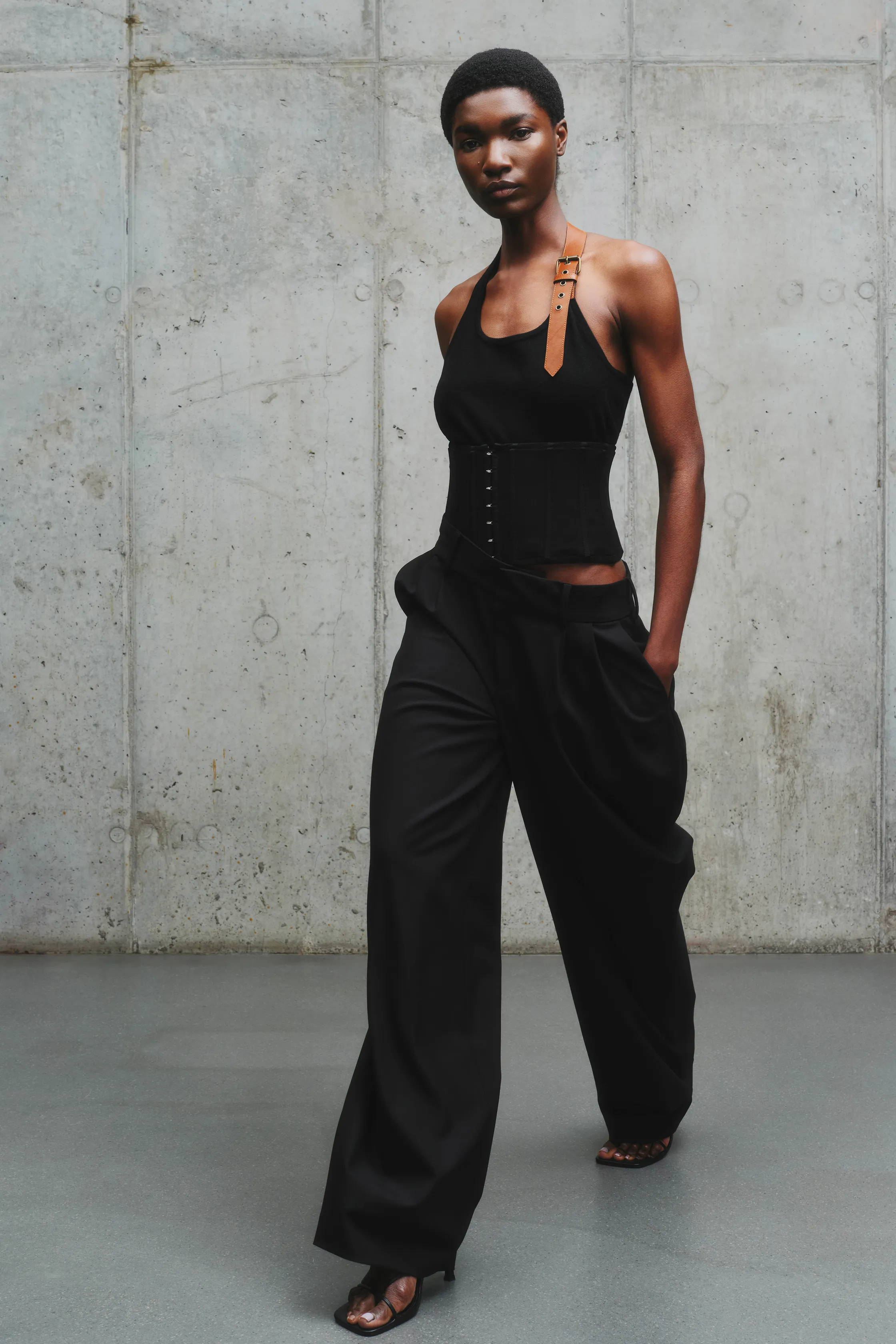 Модель в черном корсете с кожаным ремнем и широких брюках, уверенно шагает по минималистичному пространству