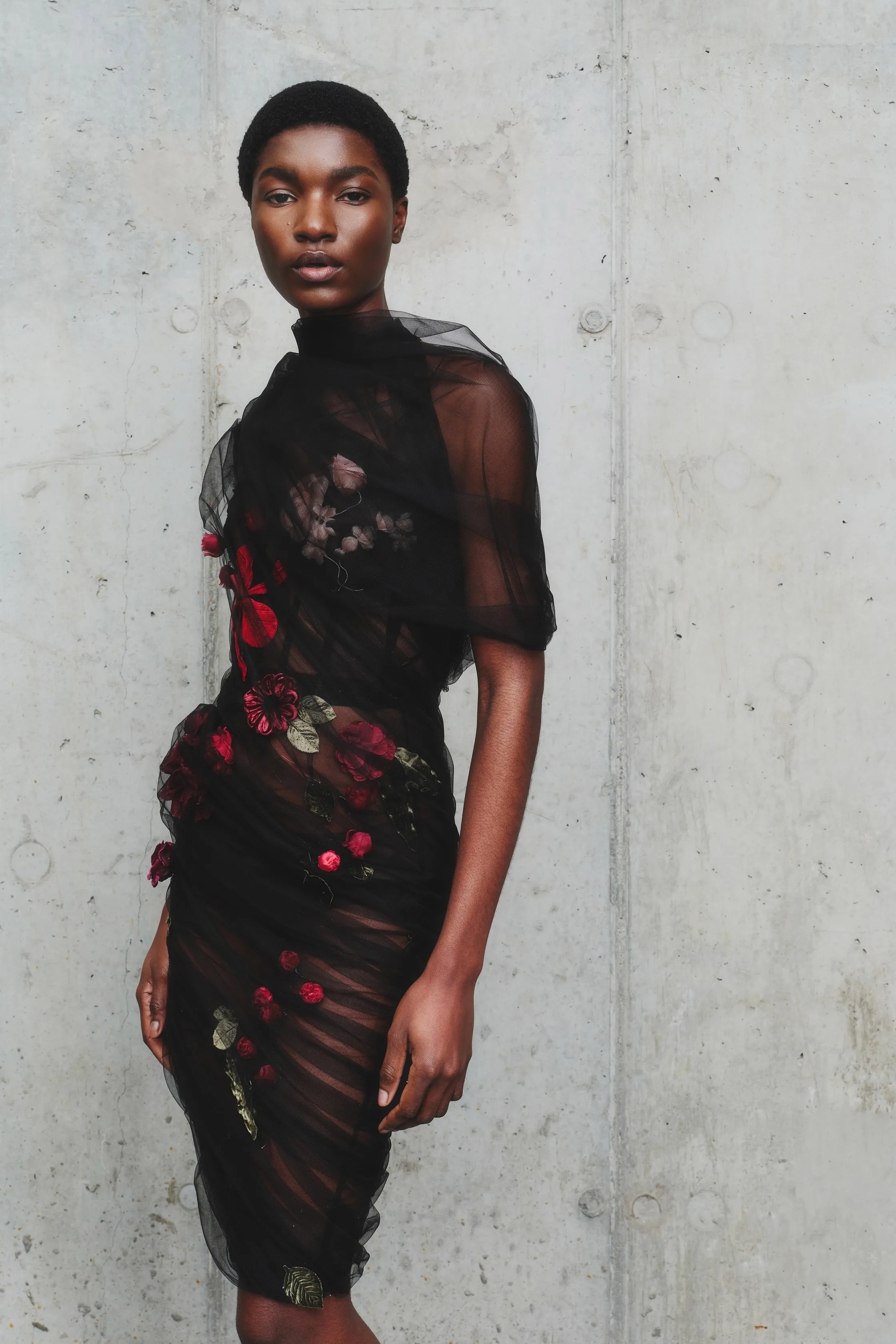 Элегантное черное платье с цветочной вышивкой, обрамляющее фигуру модели, на фоне текстурированной бетонной стены