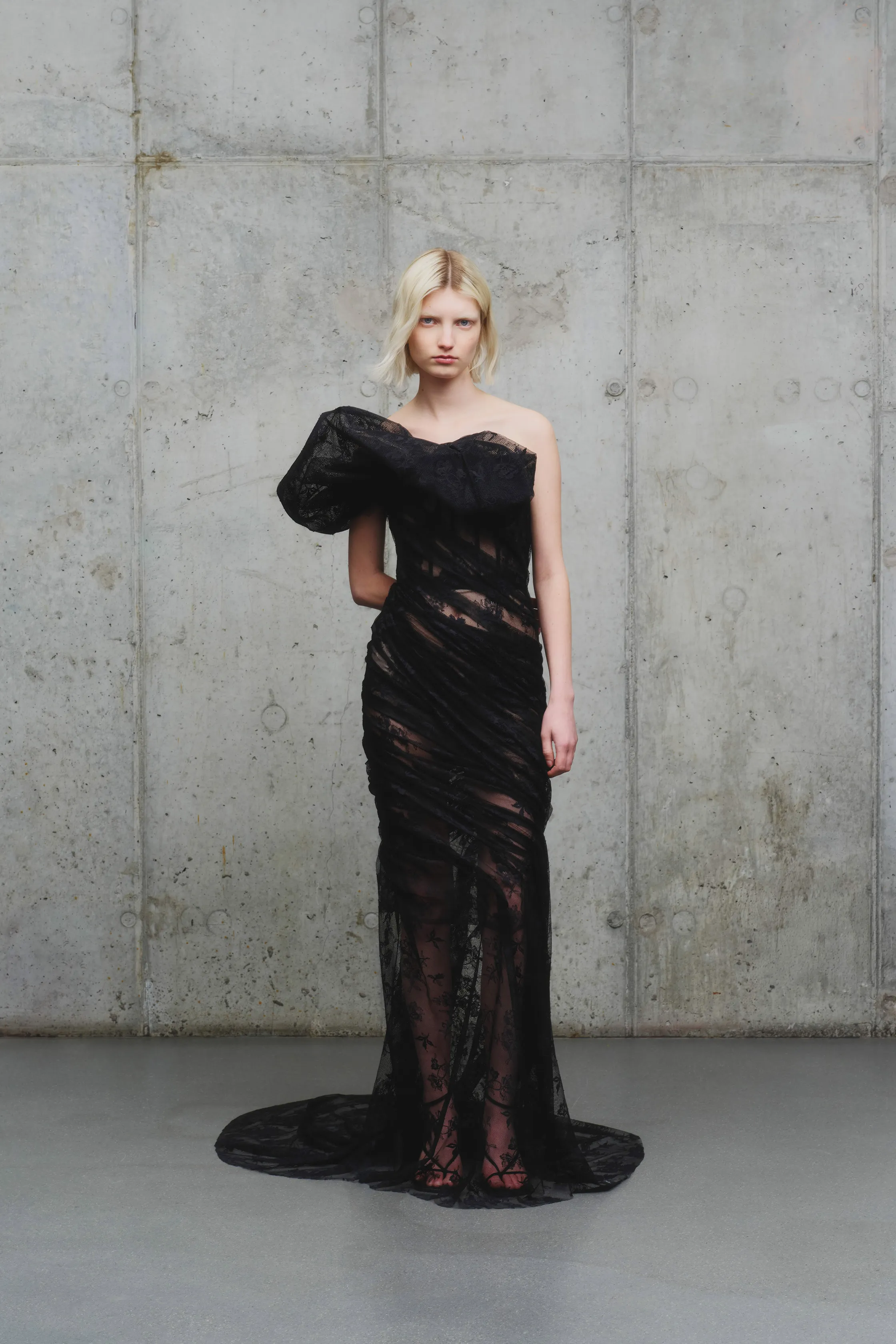 Платье с асимметричным рукавом и прозрачной текстурой, создающее эффект легкости и изысканности на фоне серого бетона