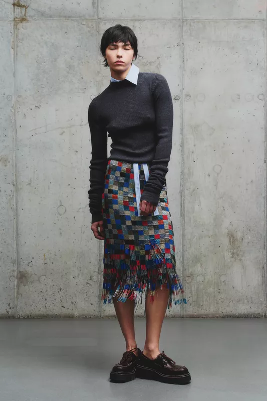 Теплый серый свитер в сочетании с яркой юбкой в клетку с бахромой, создающей динамичный образ на фоне бетонной стены