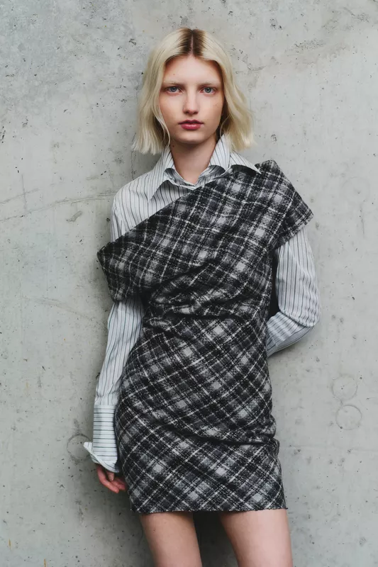 Модель в черно-серой клетчатой мини-юбке и полосатой рубашке, изящно обрамляющей фигуру, на фоне бетонной стены