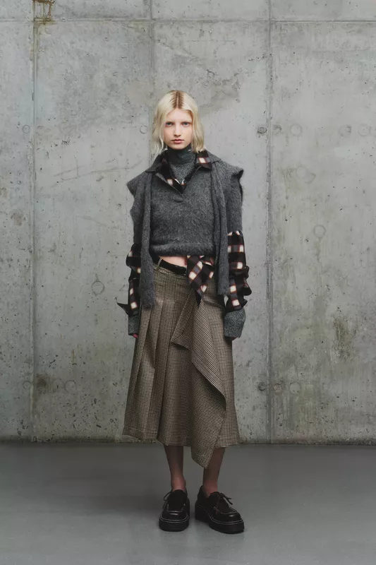 Стильный образ с серым свитером и клетчатой юбкой, дополненный многослойностью и текстурой, на фоне индустриального интерьера