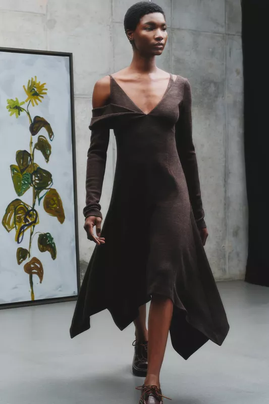 Тёмное платье с открытыми плечами и асимметричным подолом, создающее элегантный силуэт на фоне абстрактной картины