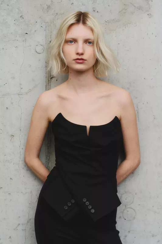 Черный топ с глубоким вырезом и асимметричным кроем, подчеркивающий стройную фигуру модели на фоне бетонной стены