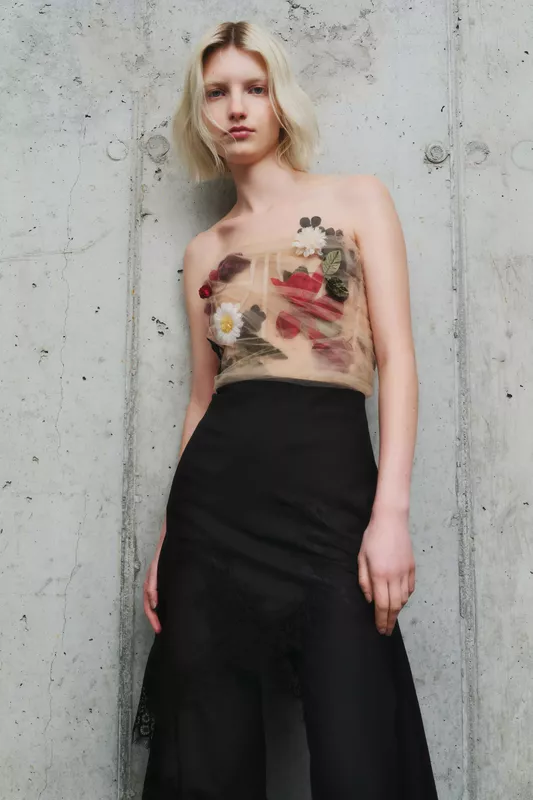 Полупрозрачный топ с цветами, контрастирующий с черной юбкой, создает романтичный образ на фоне серой стены