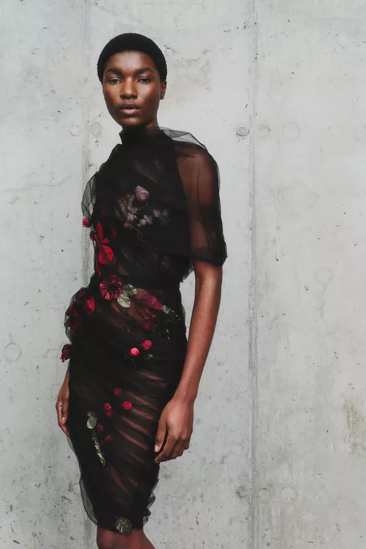 Элегантное черное платье с цветочной вышивкой, обрамляющее фигуру модели, на фоне текстурированной бетонной стены