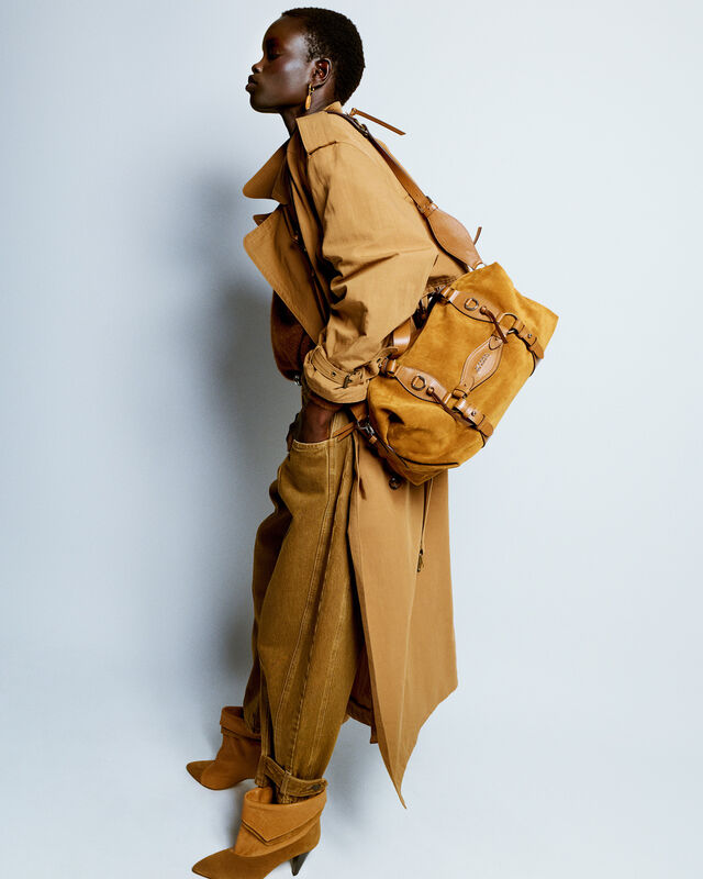 Модель в коричневом тренче и широких джинсах, дополненных стильным рюкзаком и остроносыми ботильонами, излучает уверенность