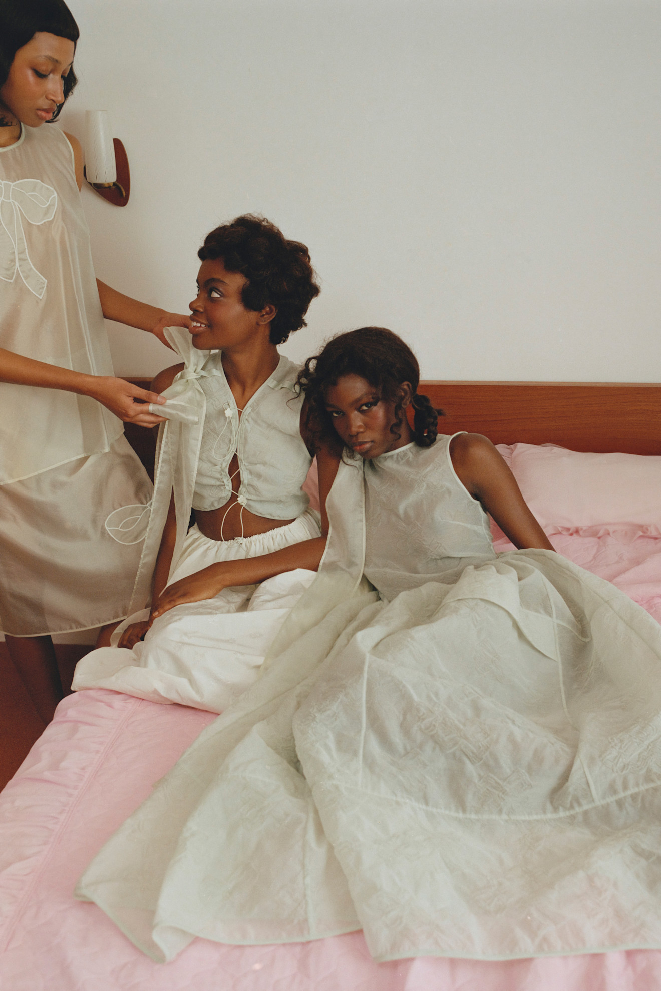 Три модели в легких нарядах из прозрачной ткани, сидящие на розовом покрывале, создают атмосферу уюта и дружбы