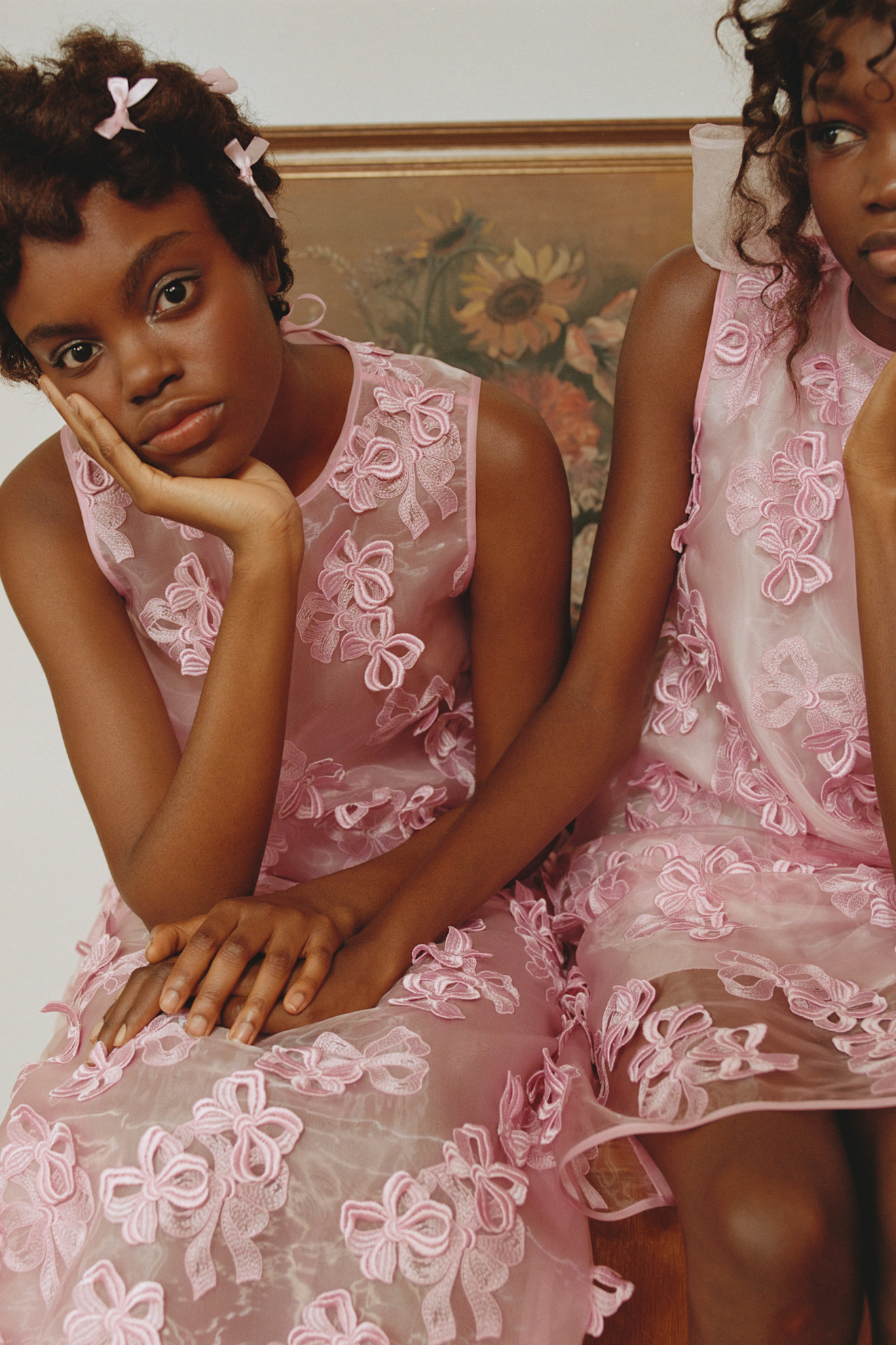 Две модели в розовых платьях с объемными бантами, задумчиво смотрят в камеру, окруженные уютной обстановкой интерьера