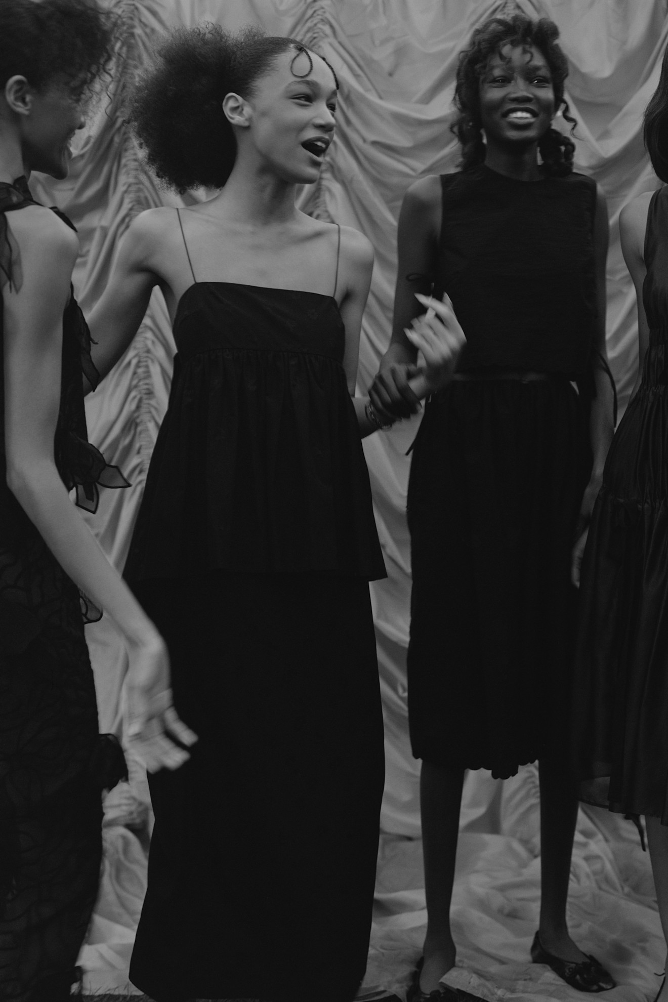 Модели в черных нарядах с легкими силуэтами, смеются и общаются на фоне драпировки, создавая атмосферу дружеского уюта