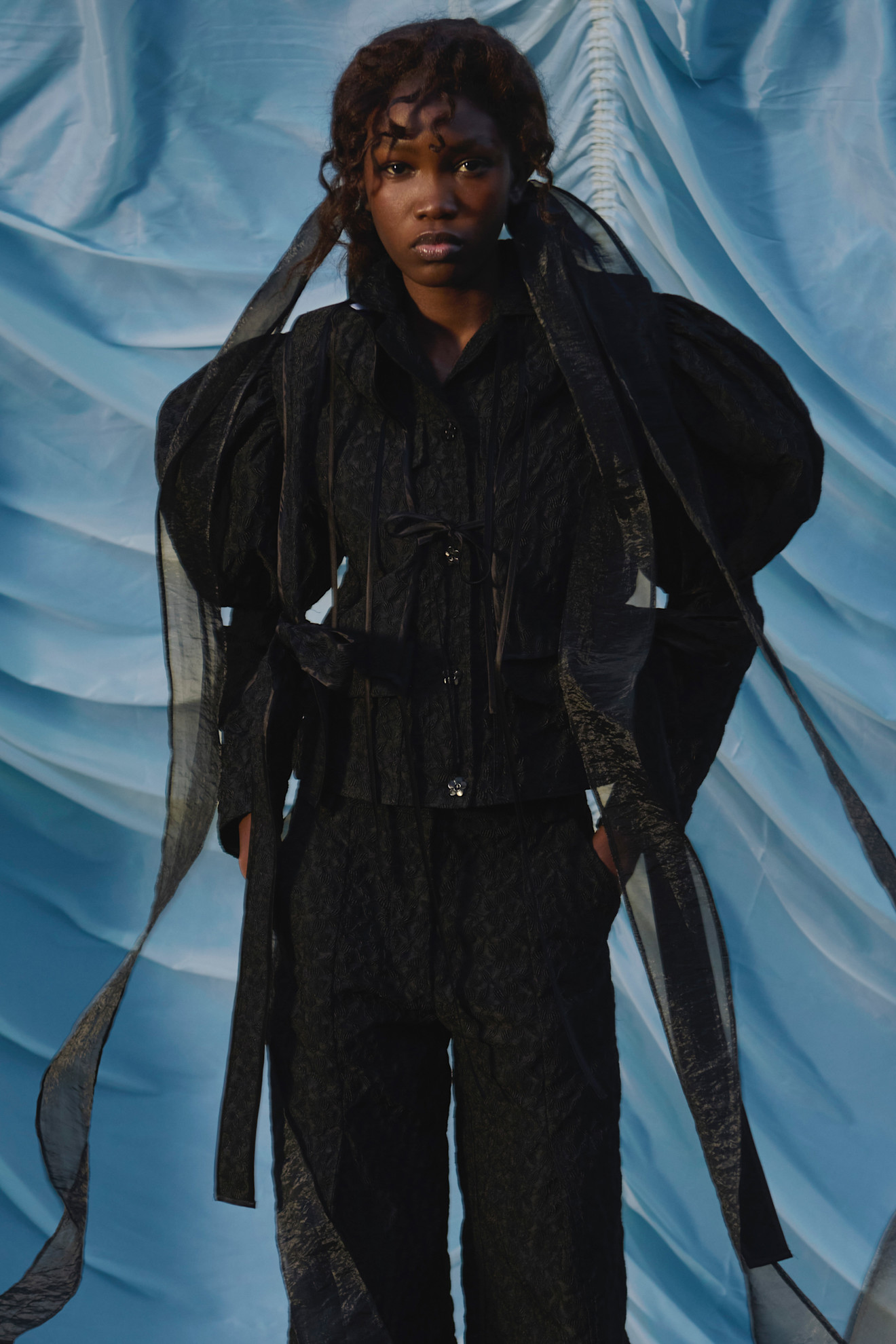 Модель в черном комплекте с объемными рукавами и прозрачными деталями, контрастирующая с ярким фоном, излучает уверенность и стиль