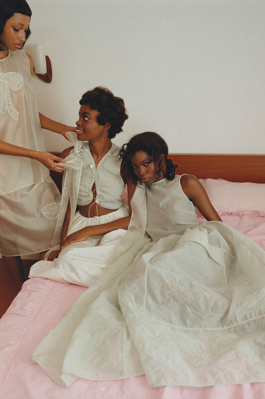 Три модели в легких нарядах из прозрачной ткани, сидящие на розовом покрывале, создают атмосферу уюта и дружбы