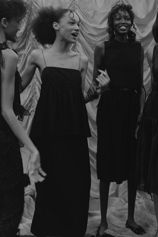 Модели в черных нарядах с легкими силуэтами, смеются и общаются на фоне драпировки, создавая атмосферу дружеского уюта