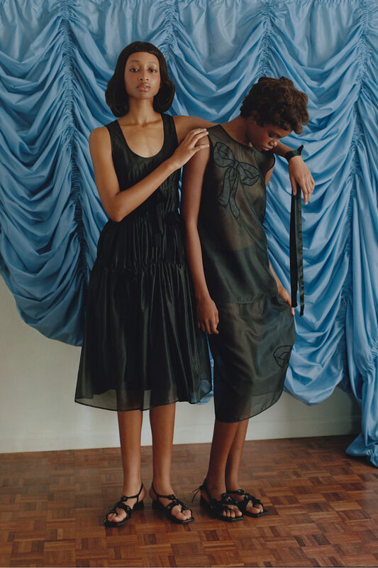 Две модели в черных платьях, одна с объемным силуэтом, другая в полупрозрачном, на фоне мягкой голубой ткани