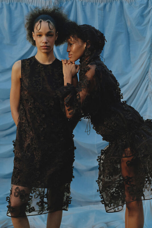 Две модели в черных нарядах с объемными цветами, позируют на фоне голубой драпировки, создавая контраст с вечерним светом