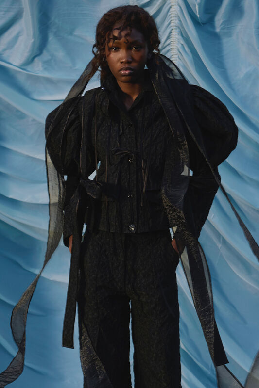 Модель в черном комплекте с объемными рукавами и прозрачными деталями, контрастирующая с ярким фоном, излучает уверенность и стиль