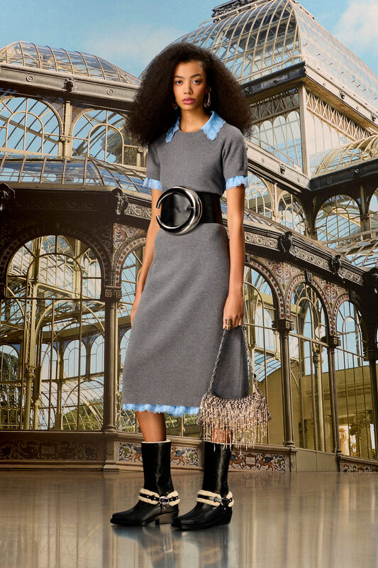 Модель в сером платье с кружевным воротником и черных сапогах, с сумкой на фоне теплицы