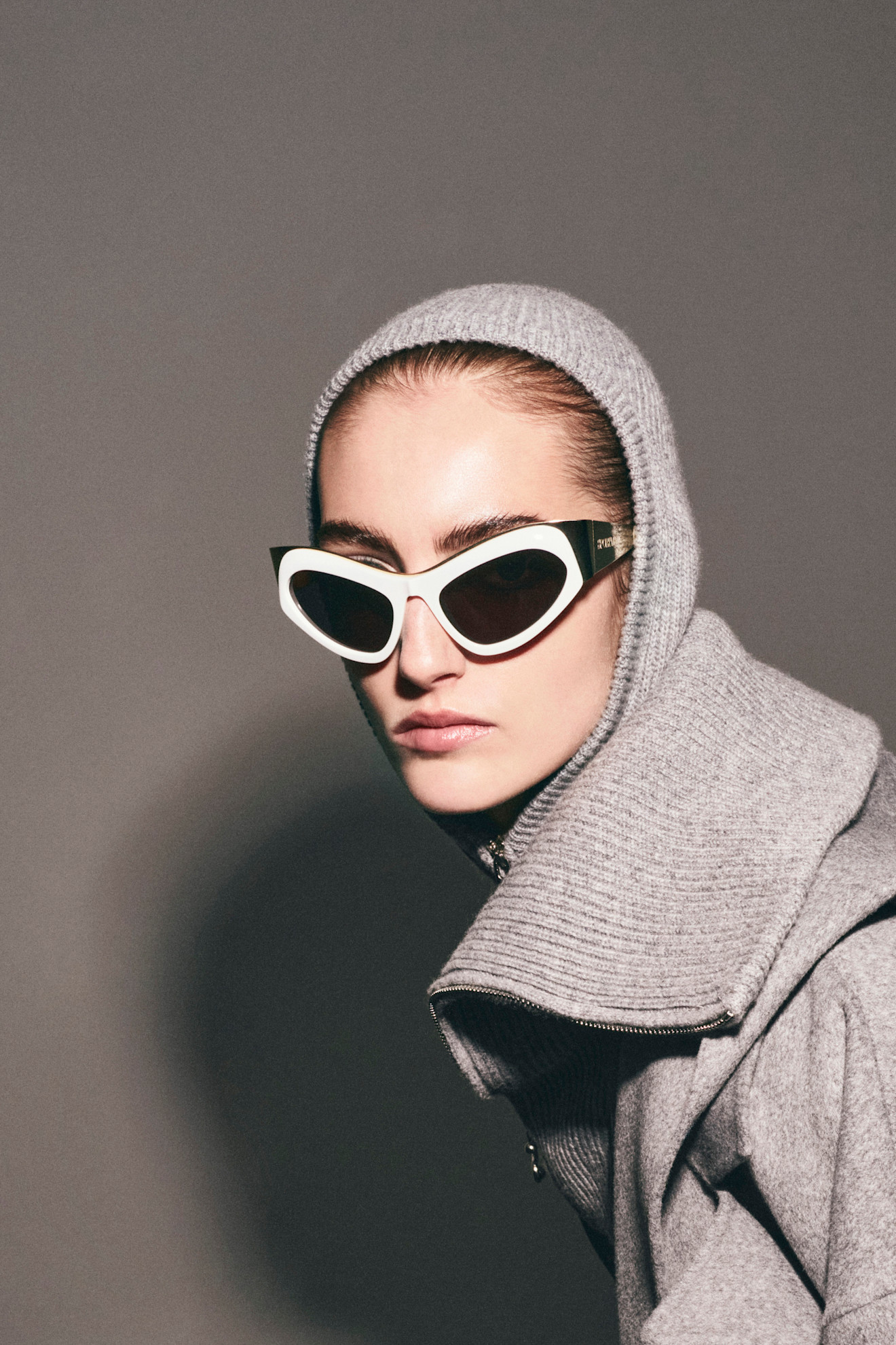 Модель в сером капюшоне и стильных очках, создающих загадочный образ на фоне нейтральной стены
