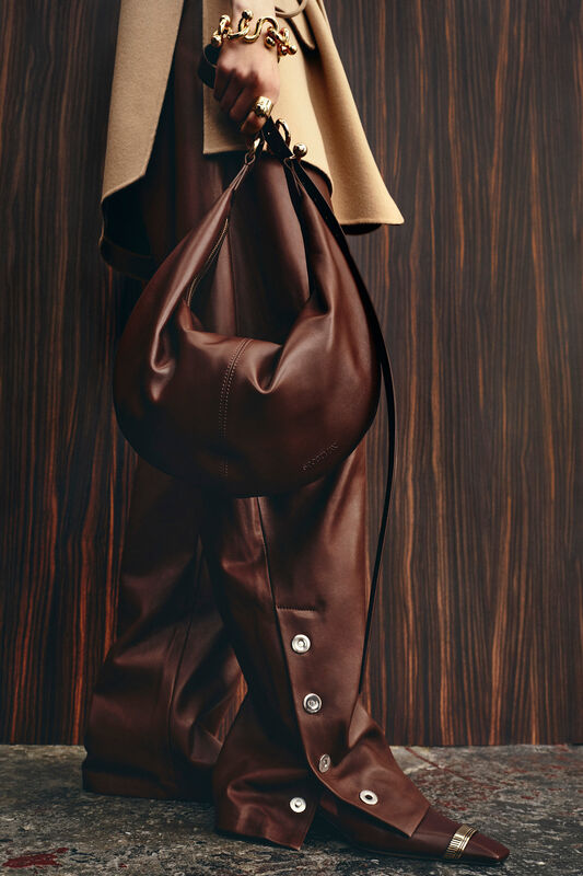 Мягкая коричневая сумка в руках, контрастирующая с бежевым жилетом и кожаными брюками, подчеркивающими текстуру