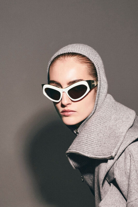 Модель в сером капюшоне и стильных очках, создающих загадочный образ на фоне нейтральной стены