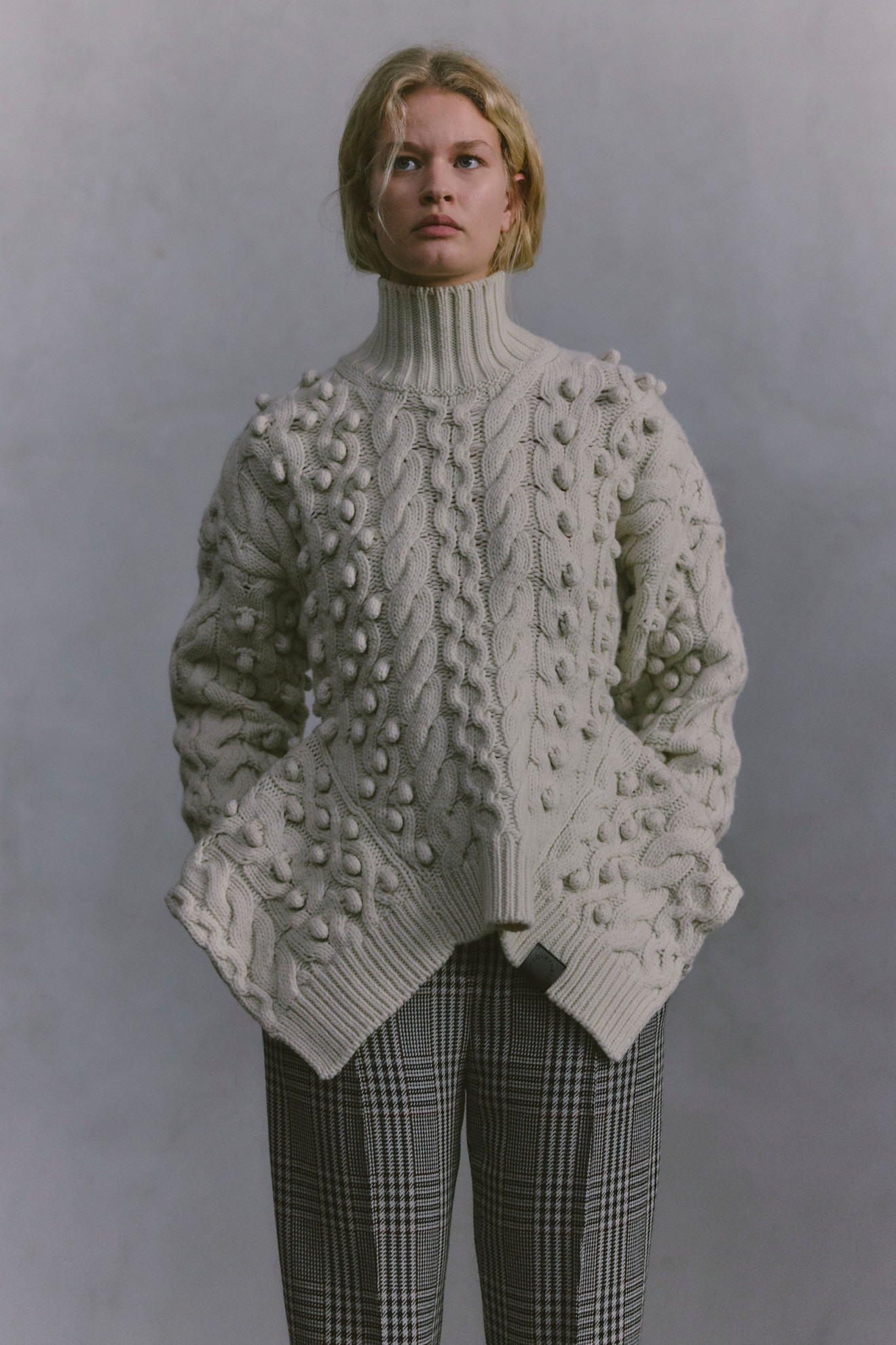 Объемный светлый свитер с текстурной вязкой и высокими воротником, дополненный клетчатыми брюками в классическом стиле